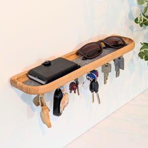 Op de afbeelding: Een houten wandplank met een grijze vilten inzet, met een zwarte portemonnee en zonnebril. Diverse sleutels hangen eronder. De plank is gemaakt van lichtgekleurd hout en is aan een witte muur bevestigd. Praktische opbergoplossing.