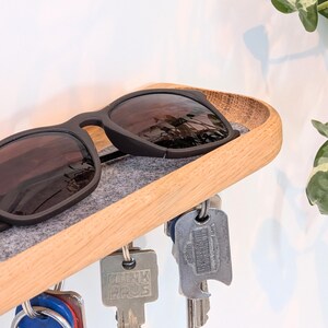 Op de afbeelding: Een houten sleutel- en zonnebrilhouder. De houder heeft een lichtbruin, gebogen houten frame met een grijze vilten inzet. Zwarte zonnebrillen rusten op het vilt. Verschillende sleutels hangen eronder.