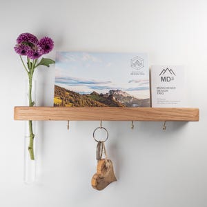 Op de afbeelding: Een houten plank met haken om sleutels aan te hangen en een kleine glazen vaas voor bloemen. De plank is gemonteerd aan een witte muur. Een ansichtkaart met een berglandschap en de tekst "Münchener Design Trio" ligt op de plank. Een klein houten hartje hangt aan een sleutelhanger.
