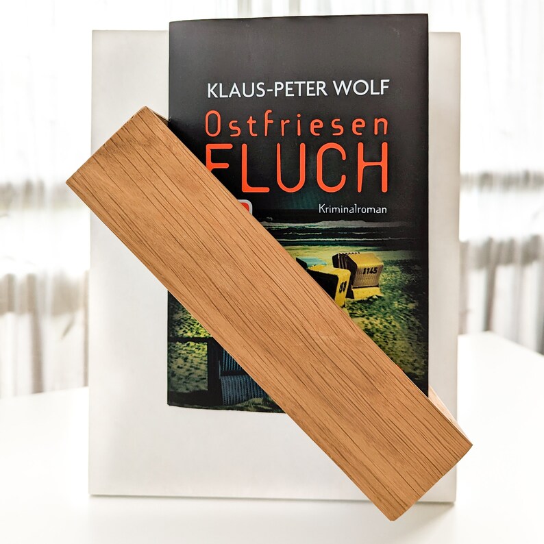 K&ouml;nnte beinhalten: Ein B&uuml;cherst&uuml;tze aus Holz mit einer nat&uuml;rlichen Holzoptik h&auml;lt ein Buch mit dem Titel "Ostfriesen Fluch" von Klaus-Peter Wolf. Der Buchumschlag zeigt eine Strandszene mit einem gelben Rettungsturm.