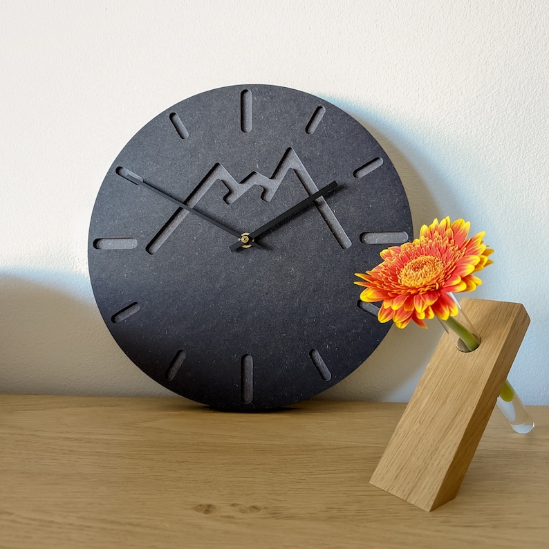 May include: A black clock with a mountain design and black hands. The clock is on a wooden surface next to a vase with a single orange flower.