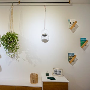 K&ouml;nnte beinhalten: Eine wei&szlig;e Wand mit einer h&auml;ngenden Pflanze in einem Makramee-Pflanztopf, einer Glaskugel, die von der Decke h&auml;ngt, und zwei Holzregalen mit B&uuml;chern darauf. Die Regale sind schr&auml;g und an der Wand befestigt. Es gibt einen Holztisch mit einem kleinen Holzkorb und einem gerahmten Foto darauf.