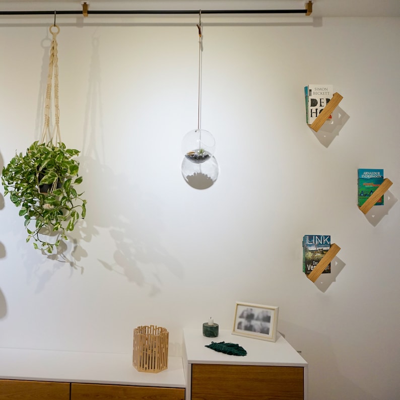 K&ouml;nnte beinhalten: Eine wei&szlig;e Wand mit einer h&auml;ngenden Pflanze in einem Makramee-Pflanztopf, einer Glaskugel, die von der Decke h&auml;ngt, und zwei Holzregalen mit B&uuml;chern darauf. Die Regale sind schr&auml;g und an der Wand befestigt. Es gibt einen Holztisch mit einem kleinen Holzkorb und einem gerahmten Foto darauf.