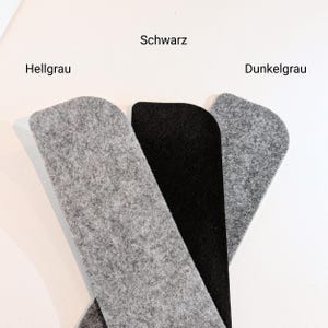 Op de afbeelding: Drie vilten stukken in grijstinten en zwart. De vilten stukken zijn gelabeld met de Duitse woorden "Hellgrau", "Schwarz" en "Dunkelgrau". De vilten stukken hebben een afgeronde bovenkant en een rechthoekige vorm.