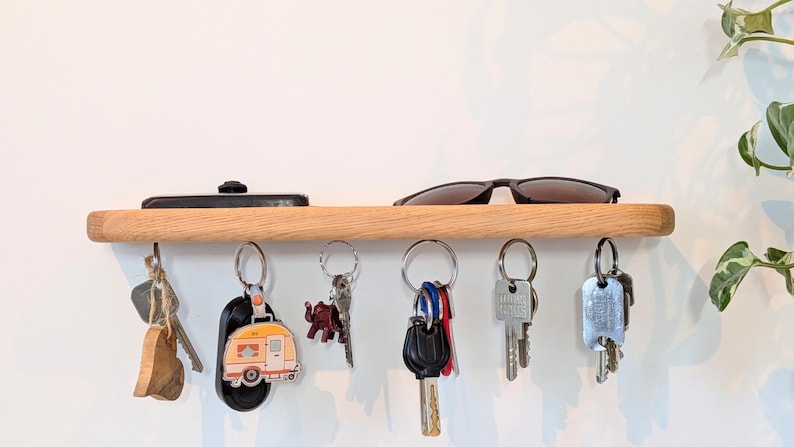 Op de afbeelding: Een houten sleutelhouder met een plank, met sleutels, een zonnebril en een telefoon. De sleutels hangen aan metalen haken. De plank is gemaakt van lichtgekleurd hout en is aan een witte muur bevestigd. Een decoratieve plant staat op de achtergrond.