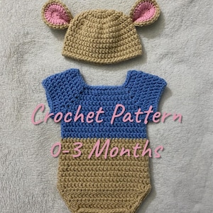 May include: A handmade crochet outfit for a baby, including a hat with pink-lined ears and a blue and tan romper. The text "Crochet Pattern 0-3 Months" is visible on the romper. The outfit is designed for newborns up to three months old.
