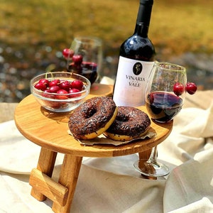Puede incluir: Una mesa de picnic de madera con dos copas de vino, una botella de vino, un cuenco de cerezas y dos donuts cubiertos de chocolate. La mesa está puesta sobre un mantel blanco.