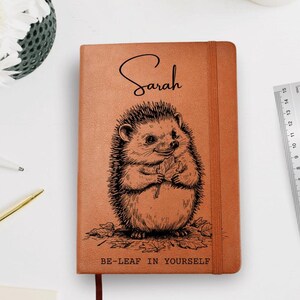 Peut inclure: Un journal marron avec le nom "Sarah" en écriture cursive. La couverture présente une illustration détaillée d'un hérisson tenant une feuille, avec le texte "BE-LEAF IN YOURSELF" en dessous. Une règle est à droite.