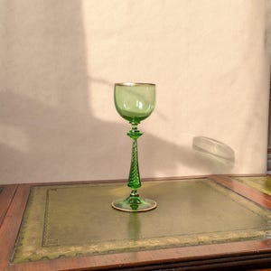 May include: A green glass goblet with a gold rim and a decorative stem. The goblet is set on a wooden surface with a green leather top. The glass has a classic, elegant design, suitable for serving wine or other beverages.