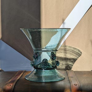 May include: A teal-colored glass vase with a flared top and a decorative base. The vase features a series of small, dark-colored embellishments around its midsection. The vase is displayed on a wooden surface.