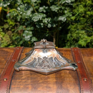 May include: An antique silver inkwell with intricate patterns and a crest design. The inkwell has a square top with a small decorative element and a wide base. It sits on a wooden surface, likely a chest or table.