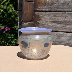 Könnte beinhalten: Eine dekorative Glasvase mit breitem, rundem Körper und ausgestelltem Rand. Die Vase weist schillernde Farben mit einem Pfauenfedermuster auf, darunter Blau- und Goldtöne. Die Vase ist etwa 13 cm hoch.