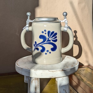 Op de afbeelding: Een beige keramische bierpul met een blauw bloemmotief en een metalen scharnierend deksel. De pul heeft twee handvatten en staat op een kleine, witte, houten kruk met drie poten. De achtergrond is een neutrale bruine kleur.