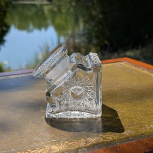 Op de afbeelding: Een heldere, zware glazen inktpot met een vierkante basis en ingewikkelde ster- en kruisarceringpatronen die in de zijkanten zijn geslepen. Het object bevat een dikke, ronde glazen stop met een getextureerde rand en een gefacetteerde bovenkant.
