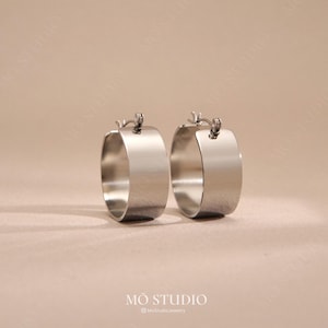 May include: A pair of silver hoop earrings. The earrings feature a thick, rectangular design with a polished finish. The image is set against a neutral background, with the brand name "MO STUDIO" at the bottom.