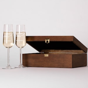 May include: Two engraved champagne flutes in a wooden box. The flutes are clear glass and have the words "Anna & Tony" etched on them. The box is brown wood with a hinged lid.