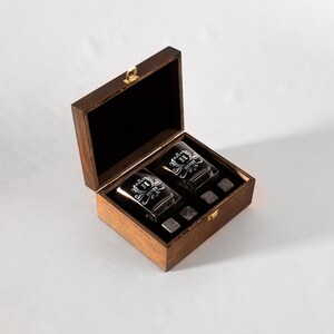 May include: A wooden box with a hinged lid, containing two engraved whiskey glasses and four whiskey stones. The glasses have a monogram and the text "Henry" etched on them.