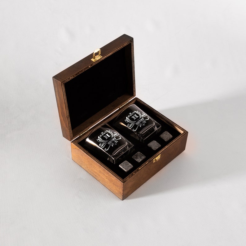 May include: A wooden box with a hinged lid, containing two engraved whiskey glasses and four whiskey stones. The glasses have a monogram and the text "Henry" etched on them.