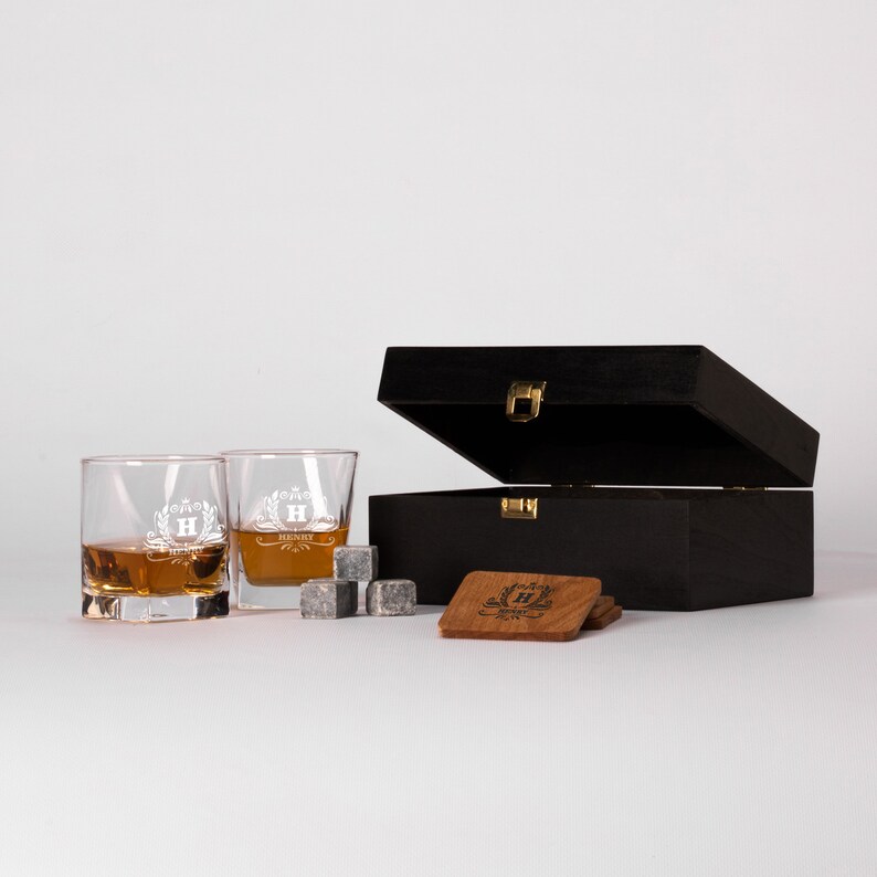 May include: A black wooden box with a hinged lid, containing two engraved whiskey glasses, four whiskey stones, and two coasters. The glasses and coasters are engraved with the letter "H" and the name "Henry".