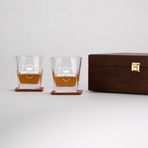 May include: Two clear glass whiskey glasses with a shield and crown design and the name "MATTHEW" etched on the front. The glasses are on wooden coasters and are in a wooden box.