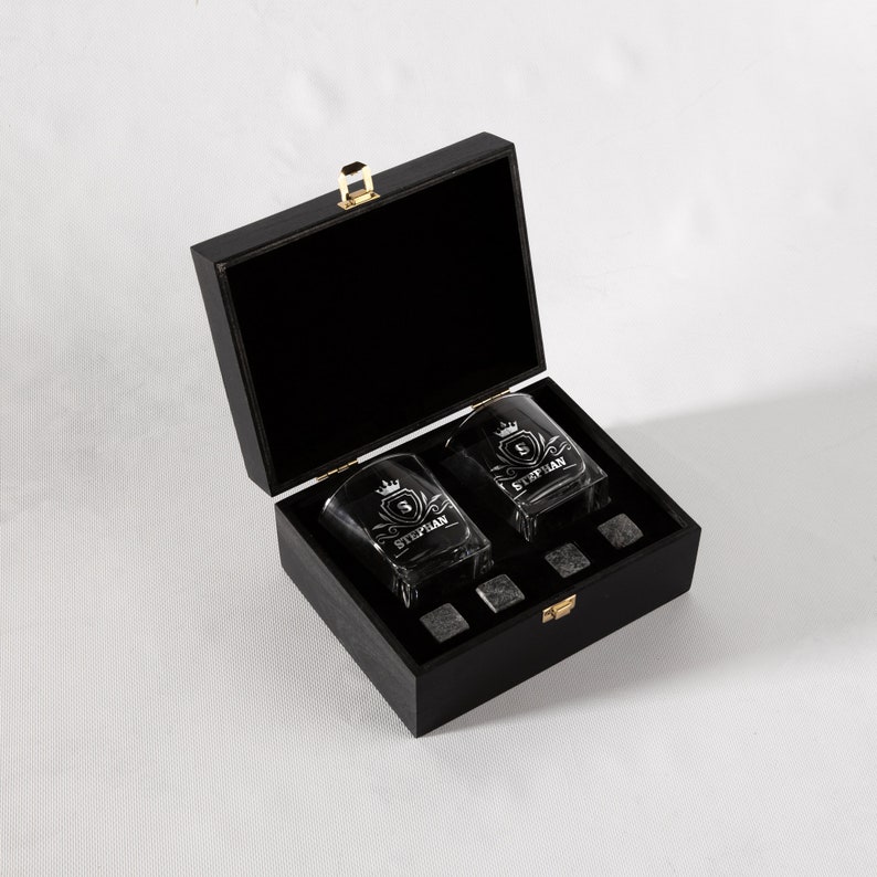May include: A black wooden box with a gold latch, containing two clear glass whiskey tumblers with a personalized inscription and a set of black stone whiskey chilling cubes.