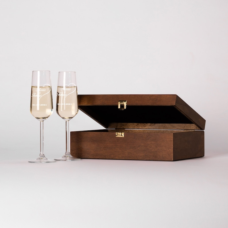 May include: Two engraved champagne flutes in a wooden box. The flutes are clear glass and have the words "Anna & Tony" etched on them. The box is brown wood with a hinged lid.