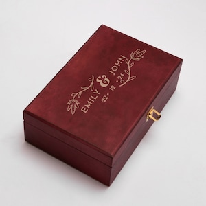 May include: A burgundy wooden box with a gold latch and engraved floral design. The box is personalized with the names "Emily & John" and the date "22. 12. 24".