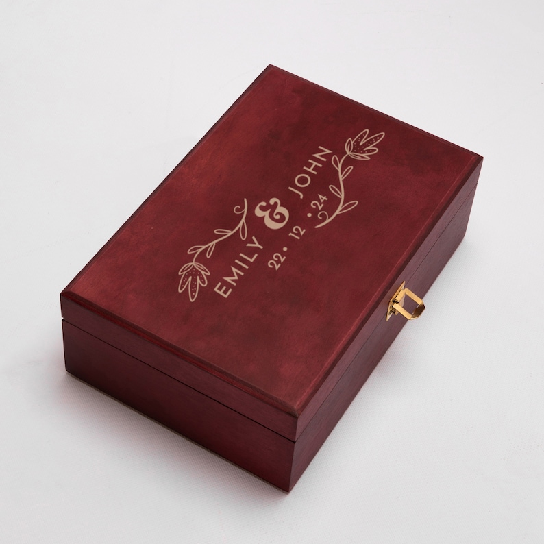 May include: A burgundy wooden box with a gold latch and engraved floral design. The box is personalized with the names "Emily & John" and the date "22. 12. 24".