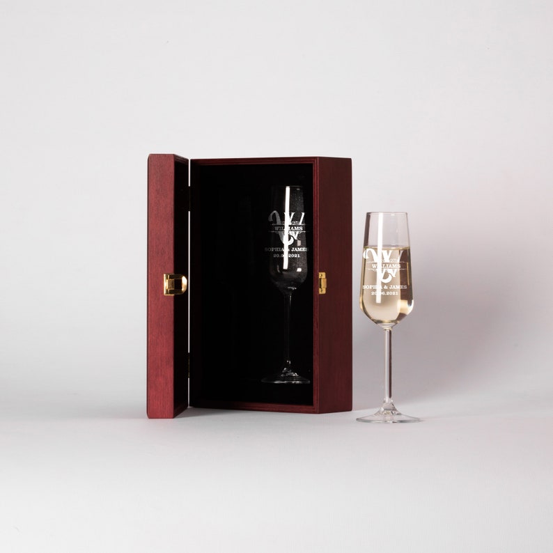 May include: A wooden box with a hinged lid is open, revealing a champagne flute inside. The flute is engraved with the text "Williams" and "Sophia & James 20.08.2021".
