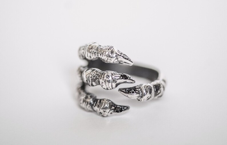 May include: A silver ring with a detailed claw design. The claws are accented with black stones.