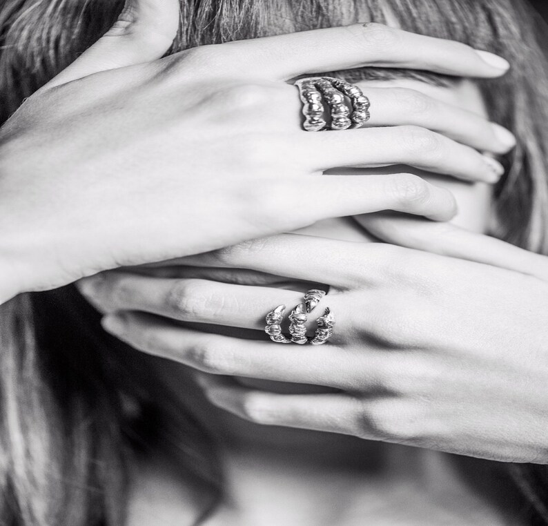 May include: Two silver rings on a person's hand. One ring is a claw design and the other is a stack of three bands.