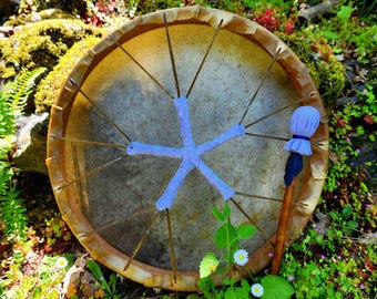May include: A brown wooden frame drum with a light blue star design and a brown wooden stick with a blue fabric head. The drum is sitting on a bed of green moss and small white flowers.