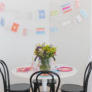 May include: A white round table is set for a party, with black chairs around it. A vase of flowers sits in the center. Decorative paper books are attached to the wall, creating a festive atmosphere.