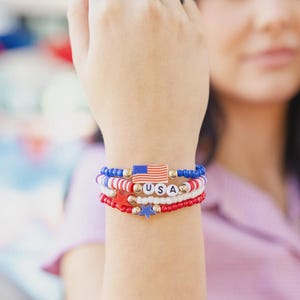 May include: A stack of beaded bracelets in red, white, and blue, with a patriotic theme. One bracelet features the word "USA" spelled out in letter beads, and another has a small American flag. Gold star and bead accents.