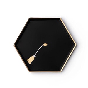 May include: A black, hexagonal tray with a gold rim and a whimsical illustration of a broom. The broom is light yellow with a white handle and a small, oval-shaped detail. The tray is set against a white background.