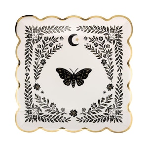 May include: A square paper plate with a scalloped gold edge, featuring a white center decorated with a black moth, a crescent moon, a small star, and a surrounding border of black floral vines and leaves printed on the surface.