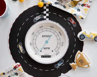 May include: A racing-themed party setting with a black and white road-shaped placemat, a plate with a speedometer design, and race car decorations. Includes napkins, cake, and a gold trophy-shaped cookie.