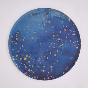 May include: A circular paper plate featuring a deep blue watercolor wash background with a scattered pattern of gold-colored dots of varying sizes across the surface, creating a celestial-inspired design on the round disposable tableware.
