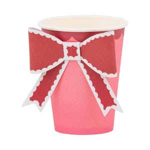 Puede incluir: Un vaso de papel rosa decorado con un lazo rojo. El lazo tiene un borde festoneado y ribete blanco. El vaso está diseñado para bebidas y es adecuado para fiestas o eventos especiales.