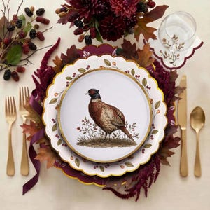 May include: A white dinner plate with a scalloped gold rim featuring a central illustration of a brown and green pheasant standing in brush, surrounded by gold-rimmed plates, gold-toned forks, a knife, a spoon, and a glass with a leaf motif.