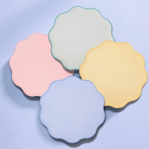 May include: A set of four circular coasters with scalloped edges in pastel colors including light pink, pale green, soft yellow, and light blue. Each coaster features a smooth, solid-colored surface with a thin, darker-toned border trim.
