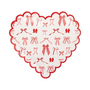 Puede incluir: Plato de papel en forma de corazón con borde festoneado rojo. La superficie blanca está decorada con varias ilustraciones de lazos rojos y rosas. Perfecto para San Valentín o una celebración romántica.