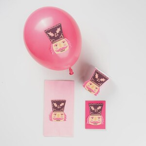 May include: A pink balloon, napkin, cup, and card with a nutcracker design. The nutcracker has a brown hat with holly, a pink face, and a yellow mustache. The items are on a white background.