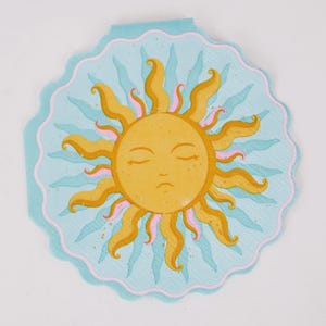 May include: A round paper napkin with a scalloped edge featuring a central yellow sun illustration with a closed-eye face. The sun has wavy golden rays with pink accents, set against a light blue background with teal wavy sunbeam patterns.