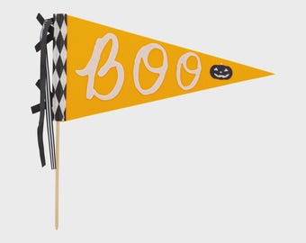 May include: A yellow pennant flag with the word "Boo" in white script, a small black pumpkin graphic, and a black and white diamond pattern. Black ribbons and a wooden stick complete the Halloween decoration.