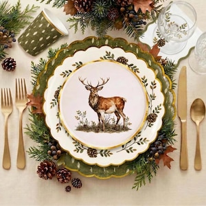 May include: A white paper plate with a scalloped gold rim featuring a brown deer illustration, layered over a larger green plate with a leaf pattern, surrounded by pine branches, pinecones, gold-toned forks, a knife, and a spoon.