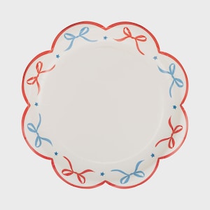 May include: A white paper plate with a scalloped edge, decorated with red and blue bows and small blue stars. The plate's border is red.