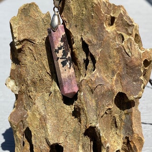 May include: A pink and black crystal pendant on a black cord. The pendant is a long, rectangular shape with a pointed end. It is hanging from a rock.