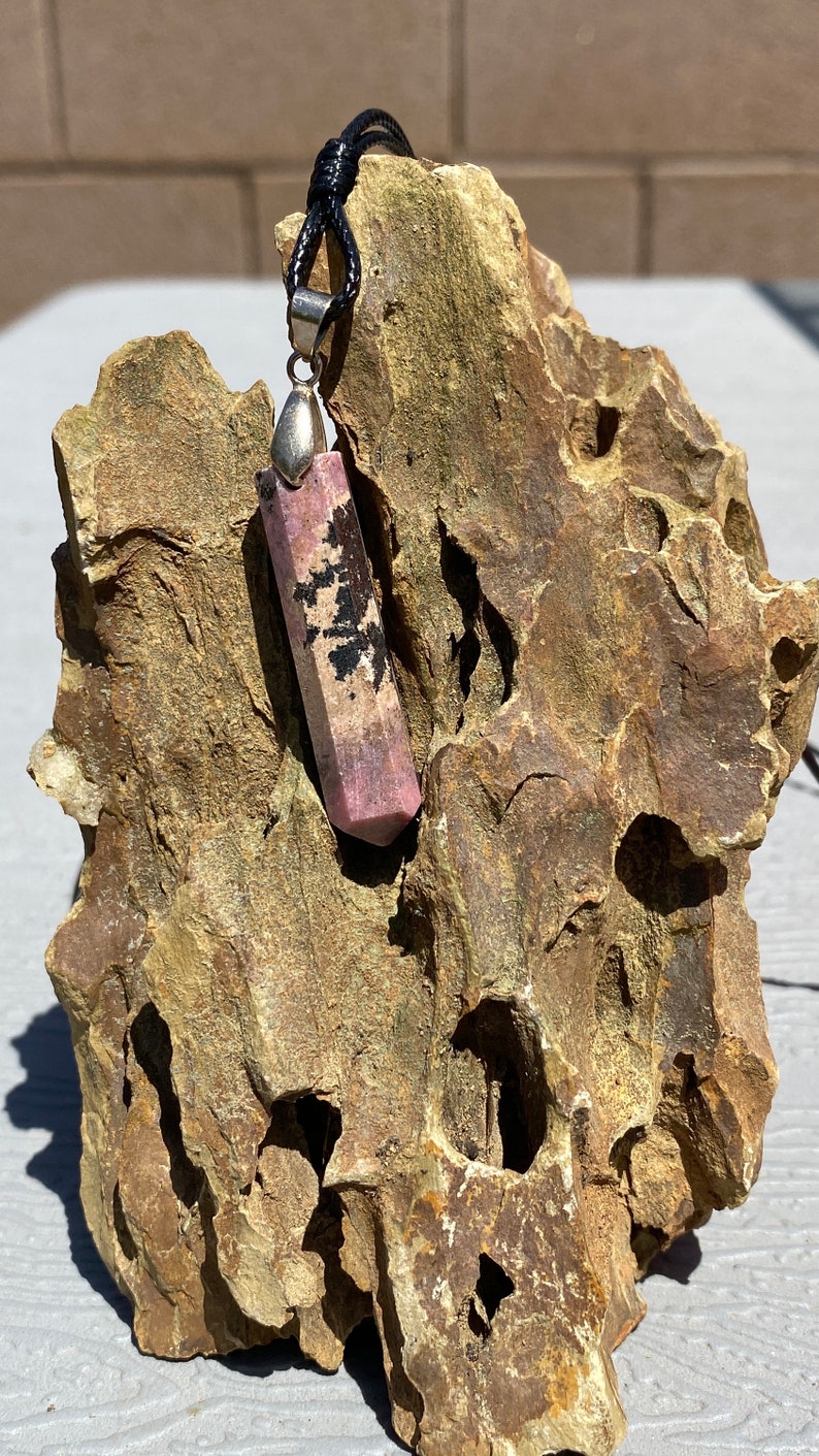 May include: A pink and black crystal pendant on a black cord. The pendant is a long, rectangular shape with a pointed end. It is hanging from a rock.