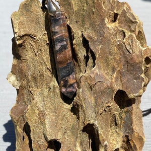 May include: A silver pendant with a long, pointed, brown and black striped crystal. The crystal is set in a silver setting and is hanging from a silver chain. The pendant is resting on a rough, brown rock.
